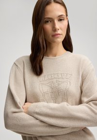 Maglione a maglia beige con scollo rotondo, caratterizzato da un emblema testurizzato sul petto. Materiale morbido e vestibilità comoda con polsini a costine.