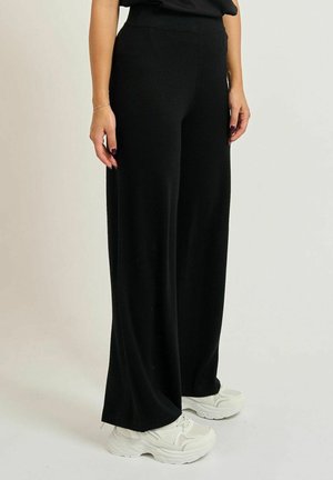 Persona che indossa pantaloni neri larghi e sneaker bianche chunky, in piedi davanti a uno sfondo chiaro uniforme.