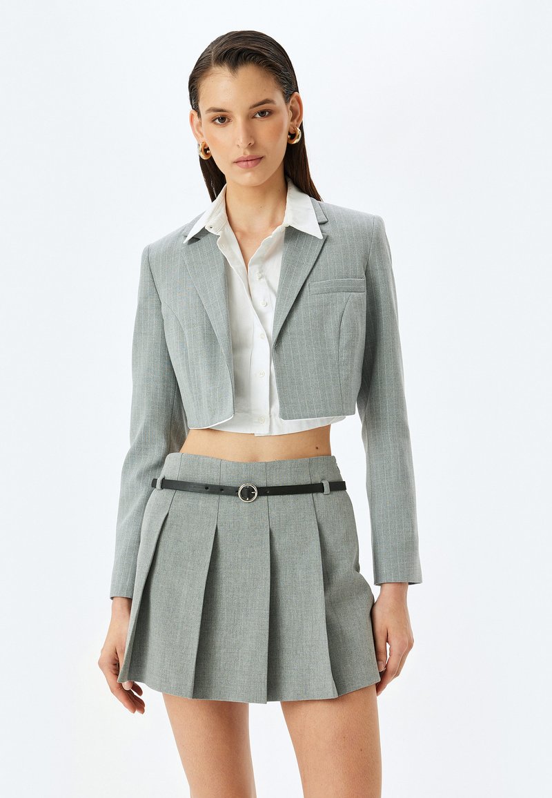 Grijs gestreepte cropped blazer over een wit shirt, gecombineerd met een grijze geplooide minirok. Zwarte riem met een ronde metalen gesp.