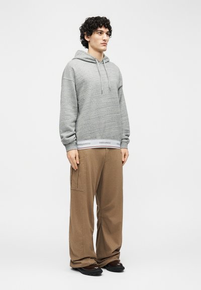 Giovane con capelli ricci che indossa una felpa con cappuccio grigia e pantaloni marroni larghi, in piedi davanti a uno sfondo bianco semplice.