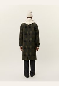 Cappotto lungo verde a quadri in misto lana, con uno spacco posteriore, indossato su un hoodie chiaro e abbinato a pantaloni scuri e un berretto.