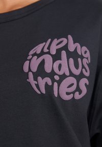 Camiseta negra con un logo burbujeante en color púrpura que dice "alpha industries", ubicado en el frente. El material parece suave y liso.