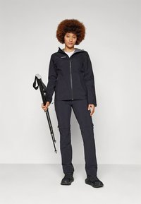 Juoda neperšlampama striukė su užtrauktuku priekyje, gobtuvu ir reguliuojamais rankogaliais, derinama su atitinkančiomis orui laidžiomis kelnėmis ir patvaria juoda avalyne.