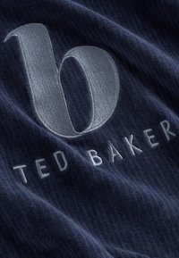 Tessuto blu navy con una texture morbida presenta una lettera "b" ricamata in grigio chiaro e il testo "TED BAKER" sottostante in un carattere elegante.