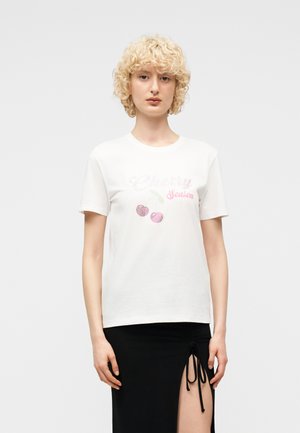 Ξανθιά γυναίκα με λευκό μπλουζάκι "Cherry Season" με ροζ κεράσια και μαύρη φούστα με άνοιγμα μπροστά δεμένη με κορδόνια.