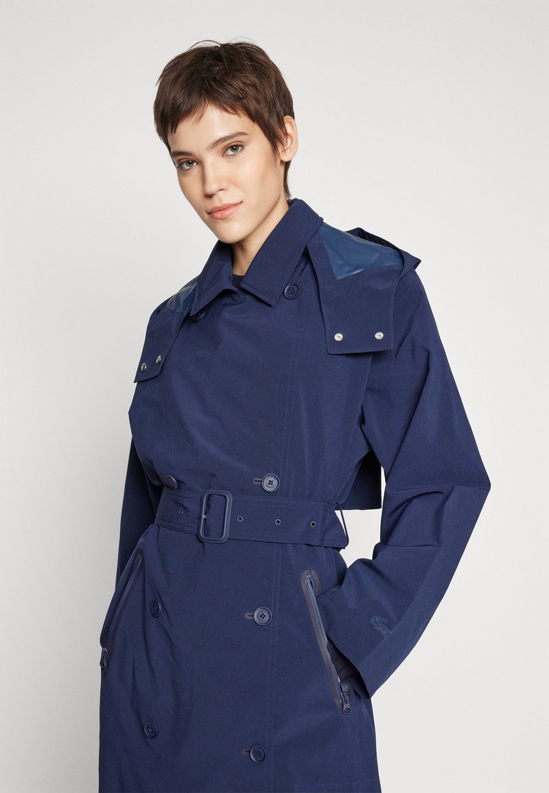 Jeune personne avec les cheveux bruns courts portant un trench-coat bleu marin ceinturé avec des poches zippées, se tenant devant un fond clair uni.