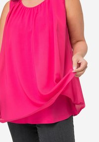 Blusa senza maniche fucsia con scollo arricciato, tessuto fluido, orlo dritto e leggera consistenza, indossata con jeans scuri.