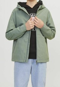 Osoba w jasnozielonej kurtce zapinanej na zamek, nosząca czarną bluzę z kapturem z logo, w połączeniu z jasnoniebieskimi dżinsami, trzymająca kurtkę przy suwaku.