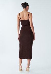 Robe midi marron sans manches avec une silhouette ajustée, une texture lisse et des bretelles fines ; portée avec des talons nus à brides.