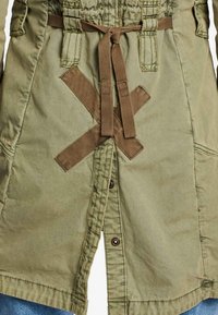 Olivgrön parkas med dragsko i midjan, som har korsstygnsdetaljer, knappar fram och sidofickor. Mjuk, vävd tyg.