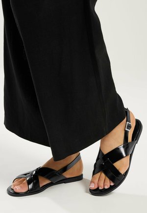 Füße in schwarzen, lackierten Sandalen mit gekreuzten Riemen und Knöchelriemen, kombiniert mit schwarzen, weit geschnittenen Hosen vor neutralem Hintergrund.