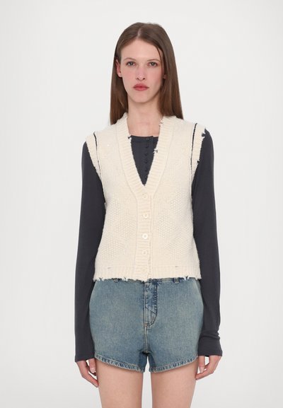 Knit cream vest with a V-neck, buttoned front, and frayed hem, paired with dark long-sleeve top and denim shorts, displayed on a neutral background.