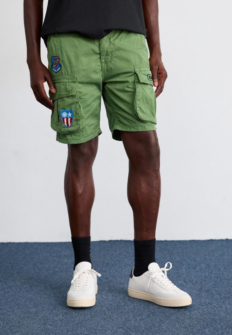 Homme portant un short cargo vert olive avec des écussons, des chaussettes noires et des baskets blanches, debout sur un tapis bleu contre un mur blanc.