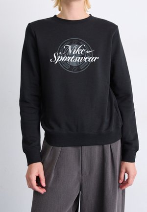 Person trägt einen schwarzen Nike Sportswear Pullover und lockere graue Hosen, steht vor einem hellgrauen Hintergrund.