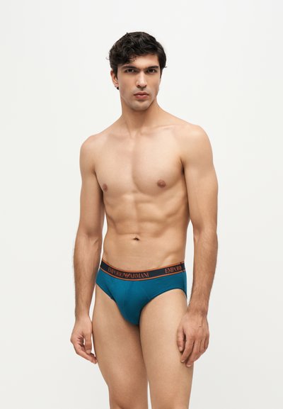 Modèle masculin aux cheveux foncés portant un slip turquoise Emporio Armani, debout devant un fond clair uni, regardant légèrement sur le côté.