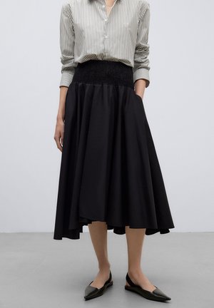 Femme portant une longue jupe noire, une chemise rayée boutonnée et des ballerines noires pointues, debout devant un fond clair uni.