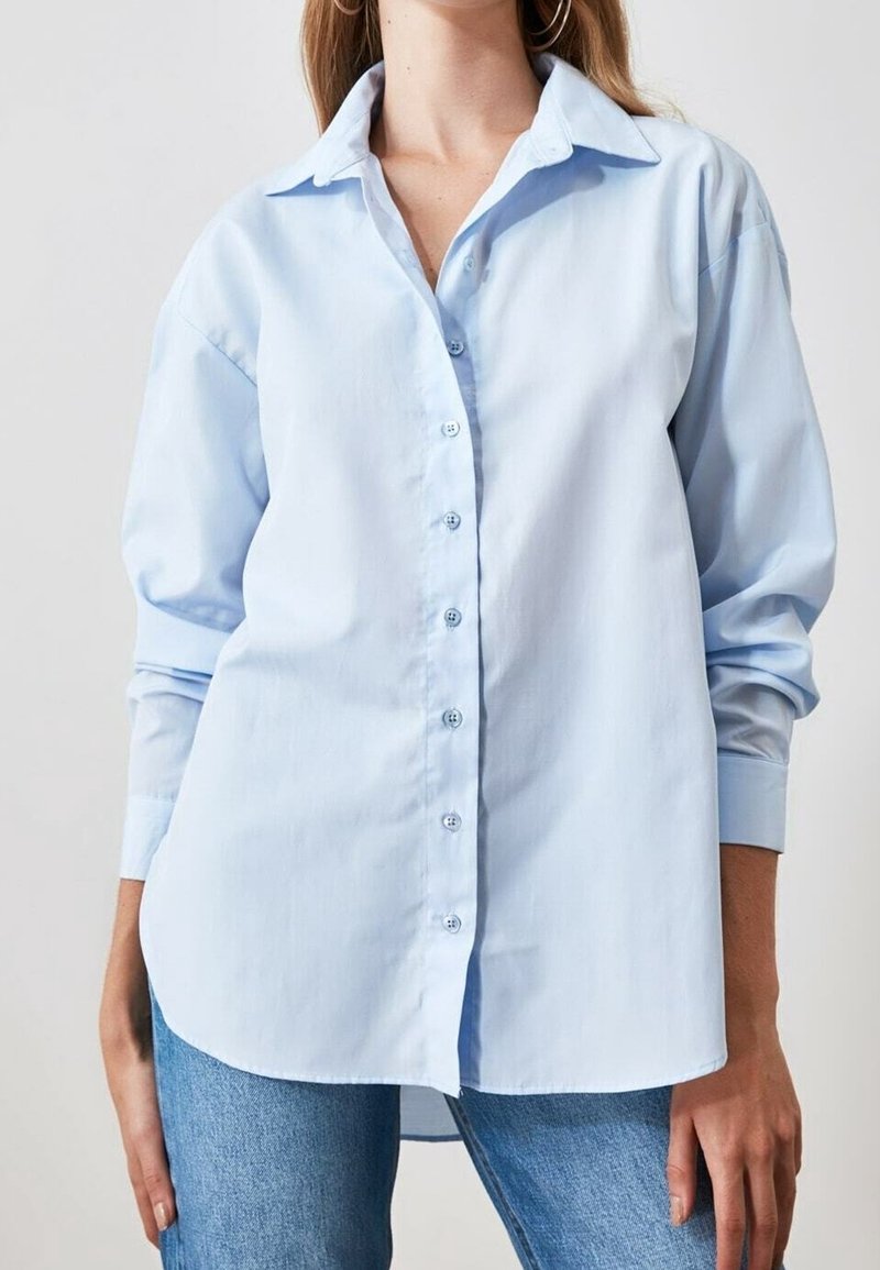 Femme portant une chemise bleu clair ample boutonnée avec manches longues et un jean bleu sur un fond uni.