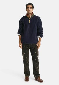 Marineblauw fleece sweatshirt met een ritskraag en groene voering, gecombineerd met zwarte corduroy broek met een vogelpatroon en bruin geknoopte schoenen.