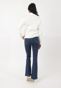 Pull en maille blanche avec un col, associé à un jean flare bleu foncé et des talons hauts de couleur claire. Design de tenue simple et décontracté.