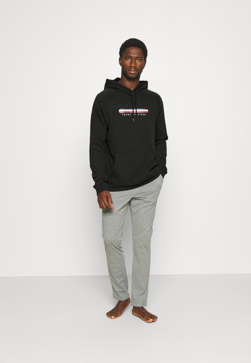 Schwarzer Kapuzenpullover mit einem weißen und roten Logo, ausgestattet mit einer Kängurutasche. Kombiniert mit hellgrauen Hosen, vor einem einfarbigen Hintergrund präsentiert.