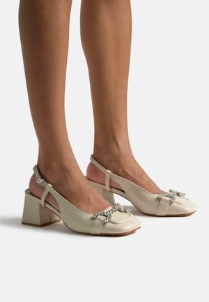 Pumps de piel beige con tirantes y puntera cuadrada, y tacón bloque. Presentan un lazo decorativo con adornos plateados.