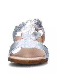 Las sandalias plateadas presentan un upper texturizado con un diseño de ondas, punta abierta y una suela plana de color marrón. Incluyen un detalle de hebilla ajustable.