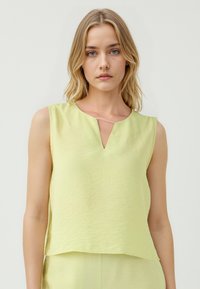 Una donna con capelli castani chiari indossa un top giallo pallido senza maniche con scollatura a V. Ha un'espressione neutra su uno sfondo bianco.