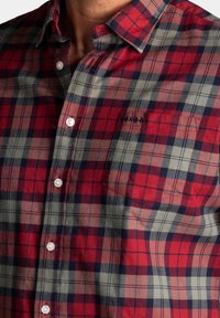 Rode, marineblauwe en grijze geruite flanellen shirt met lange mouwen en een button-down kraag. Heeft een borstzakje en witte knopen.