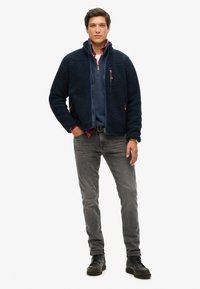 Marineblauwe fleecejas met ritssluiting aan de voorkant en zakken, grijze jeans en zwarte laarzen. Gelaagd materiaal met een casual ontwerp.