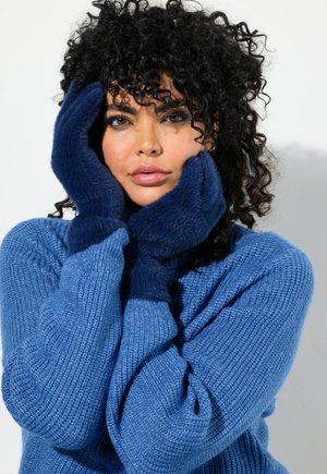 Pull en laine bleu avec texture côtelée, accompagné de gants doux bleu marine. Le mannequin pose avec les mains sur le visage contre un fond blanc.