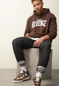 Uomo seduto su un blocco grigio che indossa un felpa marrone con la scritta "Savage Revenge", jeans neri arrotolati, calze bianche a righe e sneakers marroni.