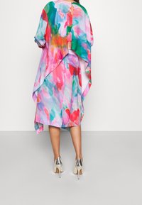 Vestido colorido y fluido en patrones abstractos de rosa, verde y azul con un diseño asimétrico. Combinado con zapatos de tacón alto plateados.