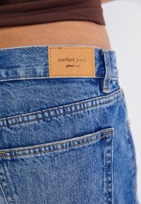 Detailní záběr na zadní kapsu modrých denimových džínů s béžovou koženou nášivkou s textem "perfect jeans gina tricot" na osobě v hnědém topu.