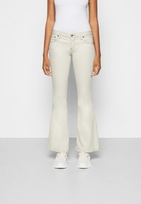 Jeans flare bege feitos de denim, com bolsos frontais, fecho de botão e uma textura suave. Usados com ténis brancos.