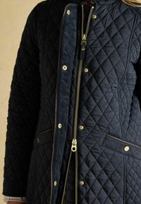 Námornícky prešívaný kabát s zlatým zipsom, patentkami a kontrastným lemovaním. Tkanina má jemný lesk a textúrovaný povrch.