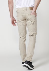 Vaqueros de algodón beige con un corte ajustado, que presentan bolsillos traseros con detalles de costura, y combinados con zapatillas negras sobre un fondo liso.