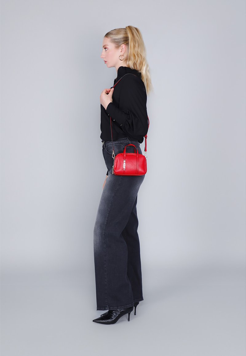 Rode leren handtas met een gestructureerde vorm en korte hengsels. Voorzien van een afneembare schouderband en zilver hardware. Model draagt een zwarte blouse en grijze jeans.