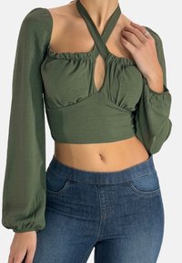 Olivengrøn, langærmet cropped top med puffede ærmer, en nøglehulsudskæring og krydsede stropper. Båret med blå, højtaljede jeans.