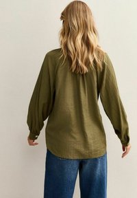 Olijfgroene blouse van textiel met lange, poffe mouwen en een knoopmanchet; de zoom valt iets lager aan de achterkant. Draag een blauwe spijkerbroek.