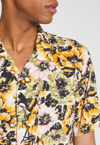Homme portant une chemise à manches courtes boutonnée avec un imprimé floral orange, jaune, noir, vert et rose et une poche poitrine.