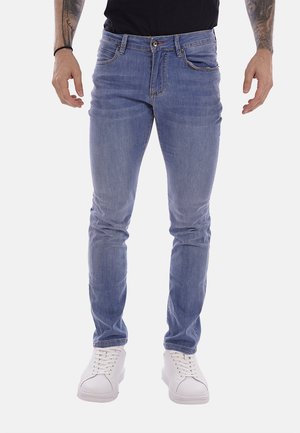 Uomo che indossa jeans slim fit azzurri chiari e sneakers bianche, in piedi con le mani rilassate lungo i fianchi contro uno sfondo semplice.