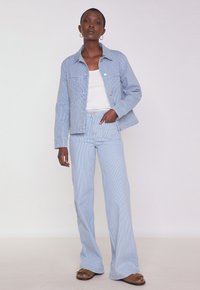 Giacca di jeans a righe blu chiaro con pantaloni a zampa coordinati. L'outfit presenta un colletto classico e dettagli delle tasche. Sotto, una semplice canotta bianca.
