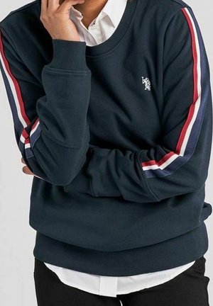 Personne portant un sweat-shirt bleu marine avec des rayures rouges, blanches et bleues sur les manches, une chemise blanche en dessous, et un pantalon noir, main touchant le visage.