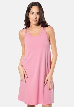 Frau in ärmellosem, knielangem rosa Kleid mit Spitzendetail an den Schultern, stehend vor einfarbig hellem Hintergrund.