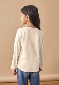 Chemise longue en ivoire clair à manches longues avec un col rond, texture douce et ourlet arrondi, associée à un jean en denim bleu, vue de dos.