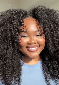 Portrett av en person med voluminøst, naturlig svart krøllete hår, iført en lyseblå skjorte, smilende under sterkt sollys.