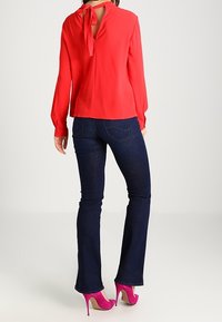 Röd långärmad blus med nyckelhål i ryggen och knytdetalj, tillsammans med mörka utsvängda jeans och livfulla rosa högklackade skor.