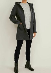 Cappotto grigio con cappuccio, design a taglio dritto, chiusura con zip, due grandi tasche, indossato sopra un maglione grigio chiaro e pantaloni neri, abbinato a stivaletti verdi.