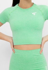 Žena v zeleném bezšvovém krátkém topu s krátkým rukávem a legínách s vysokým pasem, s rukama položenýma na bocích na bílém pozadí.