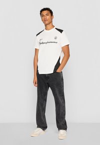 White t-shirt with black accents, featuring a logo and the word "Performance." Paired with dark, loose-fitting jeans and white sneakers.